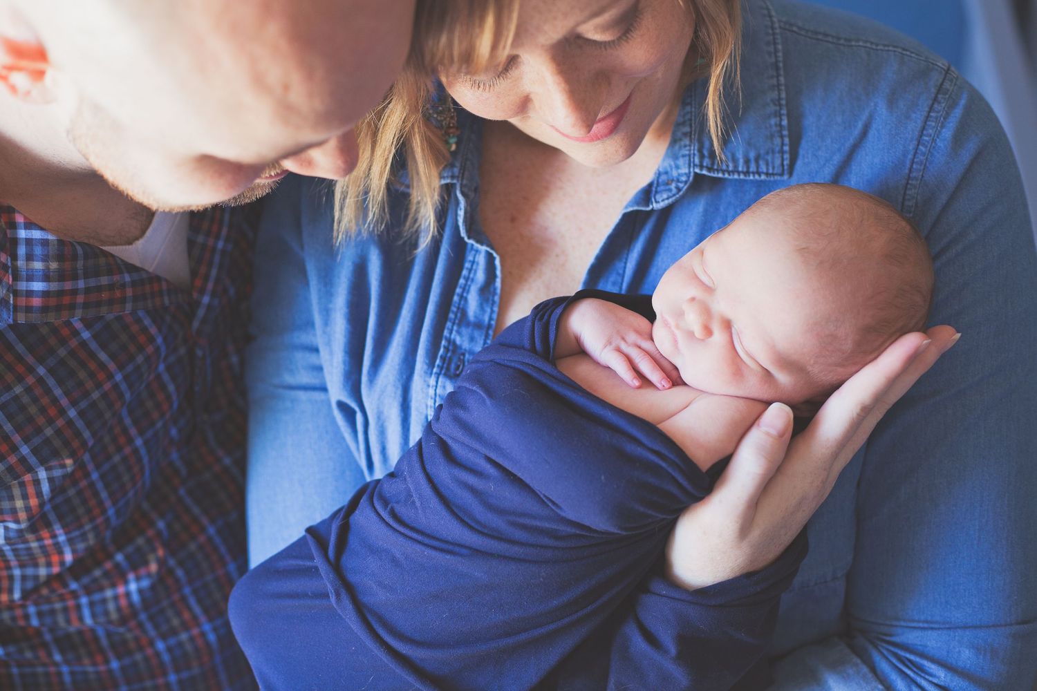 A quiet family moment as parents gaze at their sleeping newborn wrapped in blue.
