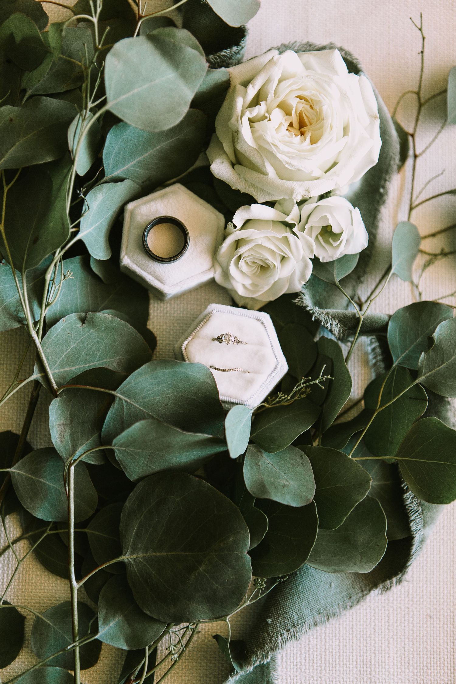 Wedding rings and white roses styled with eucalyptus leaves on light fabric.