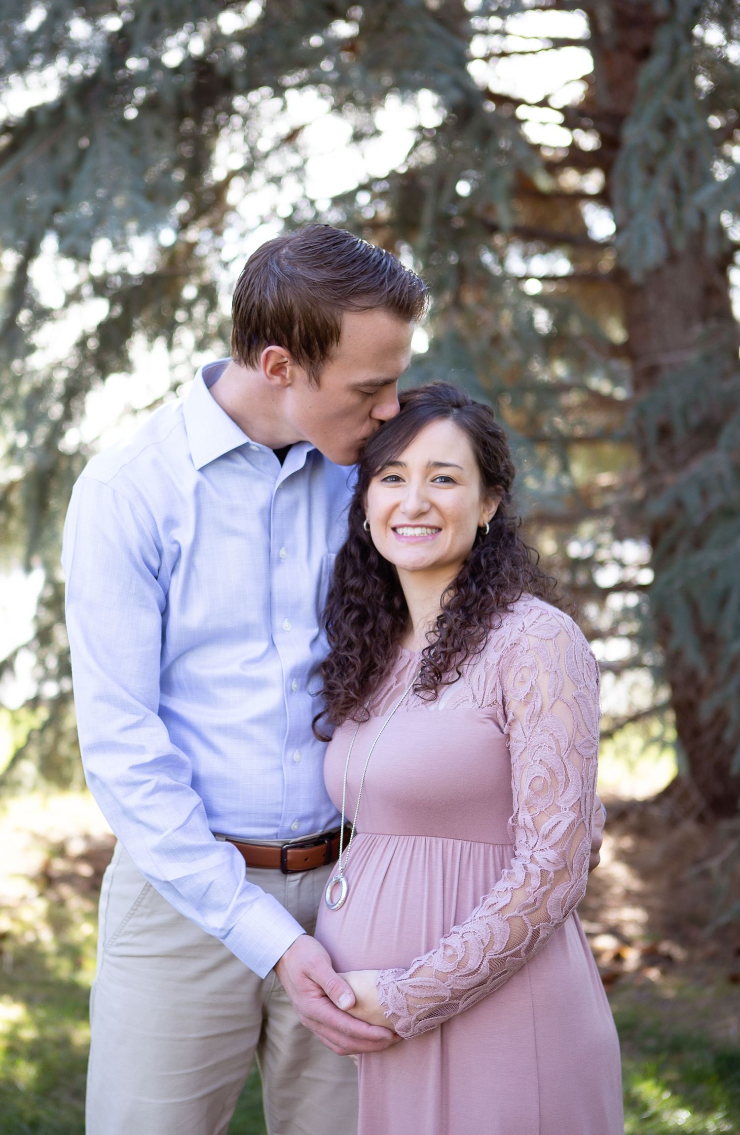 A romantic outdoor maternity photo of a couple embracing near evergreen trees.