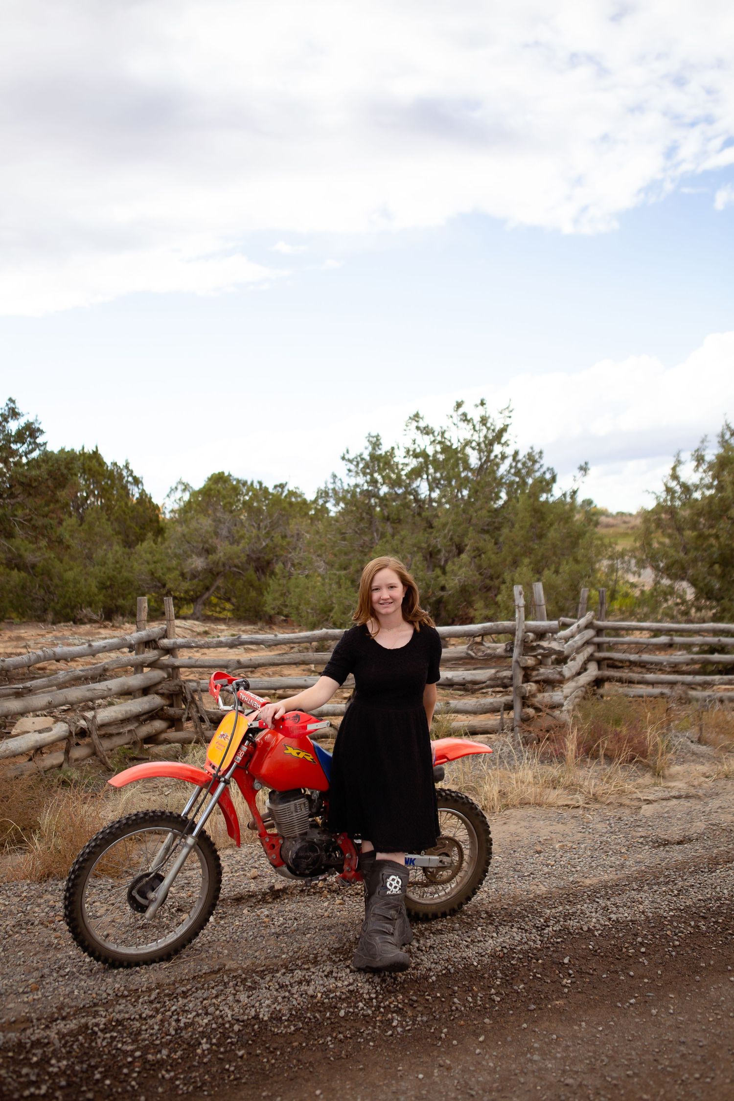 A person in a black dress poses with a red dirt bike on a gravel path near a wooden fence.