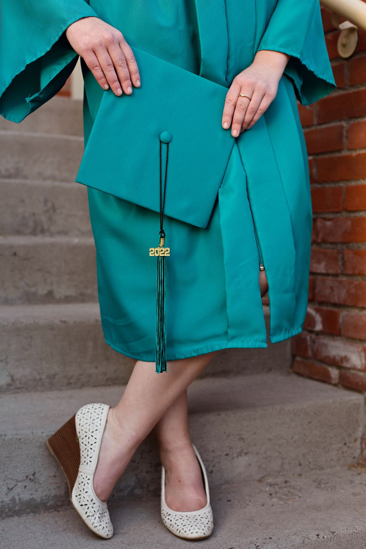 Close-up of teal graduation gown and white lace flats on stone steps with brick wall background.