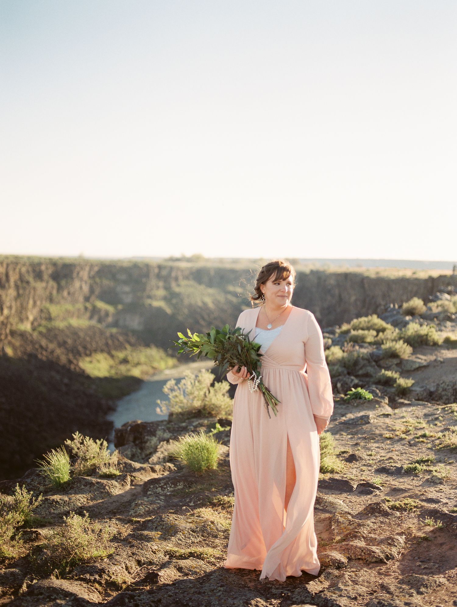 A woman in a flowing pink dress holding a bouquet stands on a cliff overlooking a scenic canyon view.