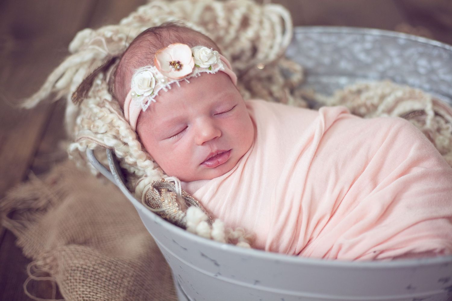 A sleeping newborn in pink wrap nestled in a vintage metal basin with lace accents.