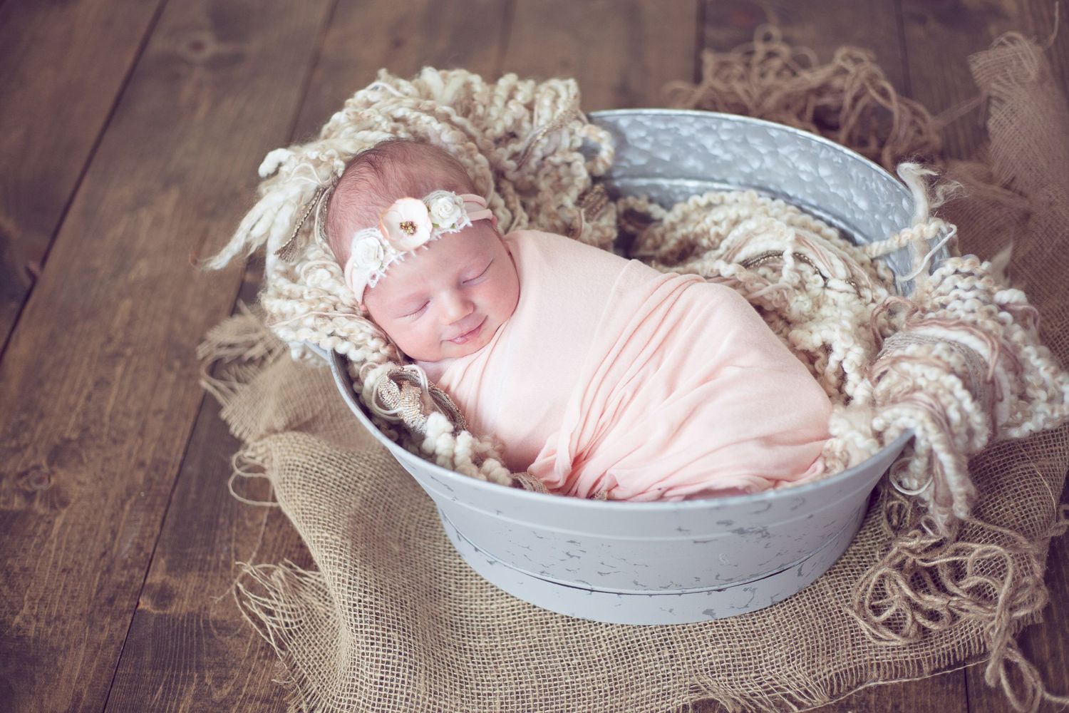 A peaceful newborn sleeping in a rustic metal basin surrounded by cream colored textiles.