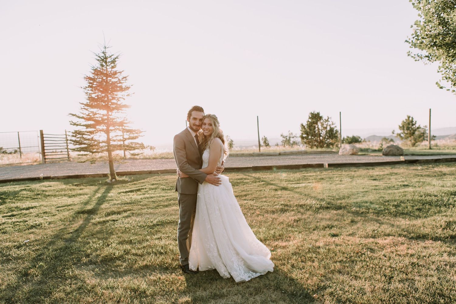 Sunset wedding portrait of couple embracing on grassy field with evergreen trees in background.