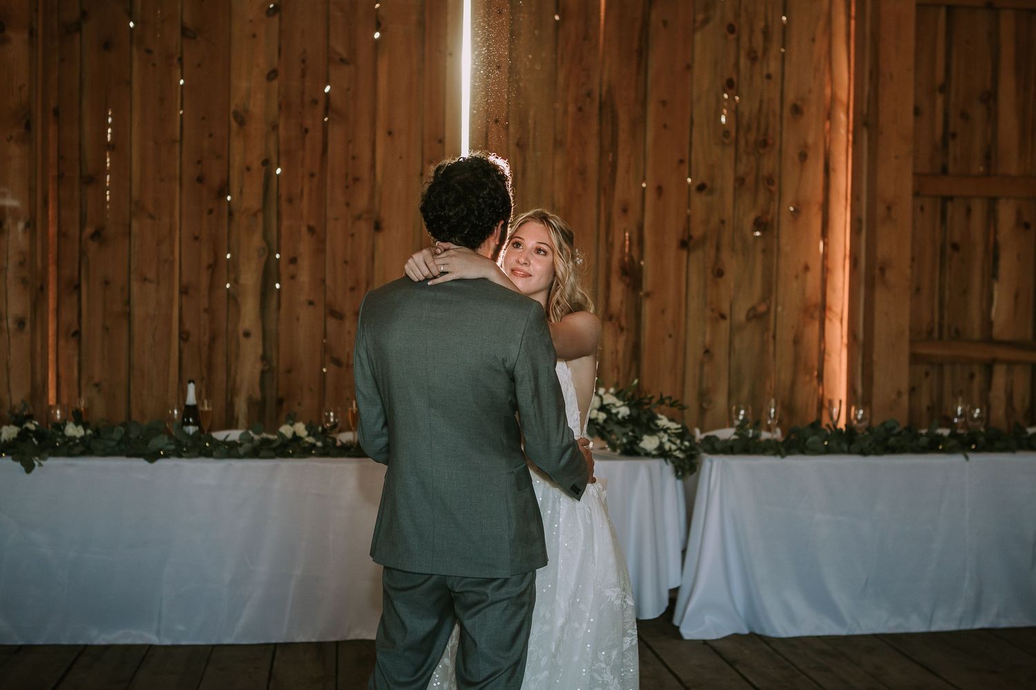 Couple embracing during their wedding dance in a wooden barn with white decorated tables.