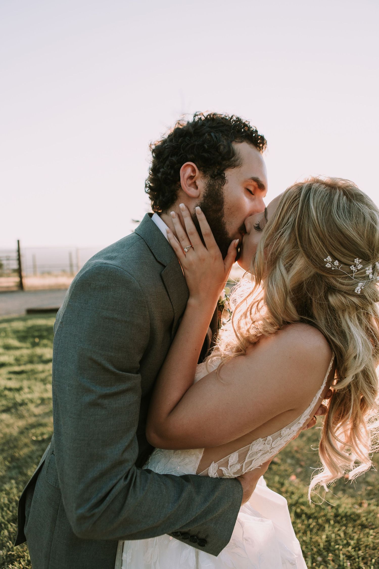 Newlyweds share a romantic kiss in formal attire against a sunset backdrop.