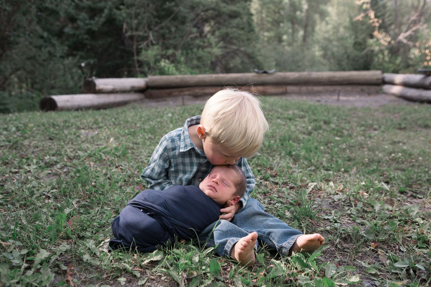 Two siblings playing together on grassy ground in a wooded area.