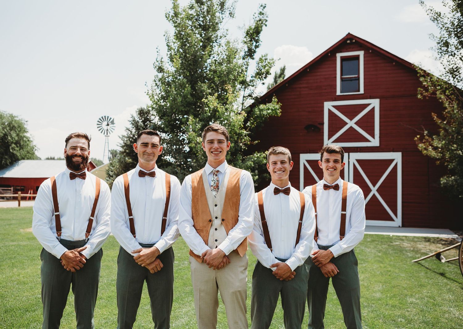Wedding party poses in front of a red barn wearing suspenders and casual attire.