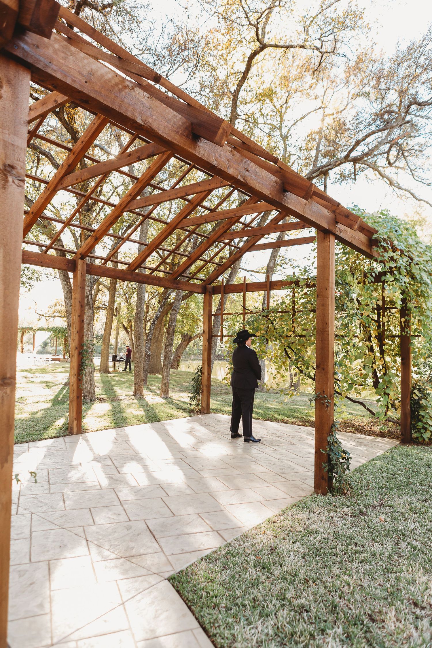 Silhouette stands in rustic wooden pergola during golden hour at wedding venue.