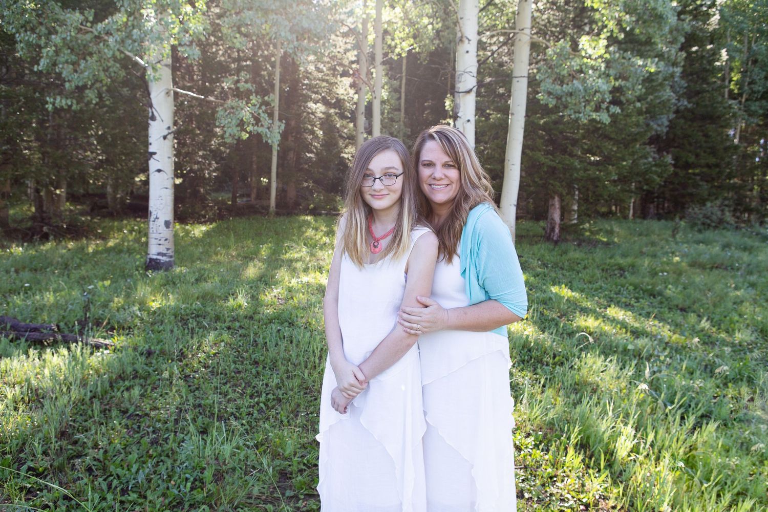 Two figures in white dresses stand together in sunlit forest clearing with aspen trees.