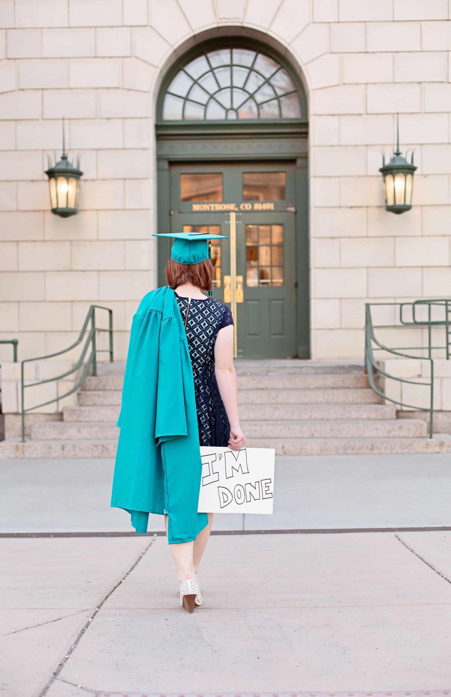 Graduate in teal cap and gown walking towards ornate building entrance holding 'I'm Done' sign.