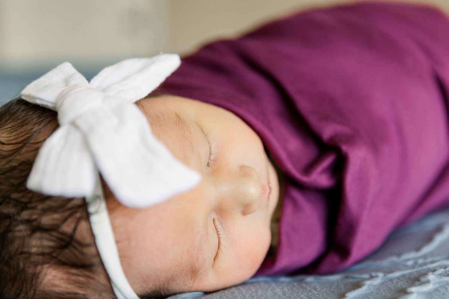 Peaceful sleeping baby in purple wrap and white bow headband on gray bedding.