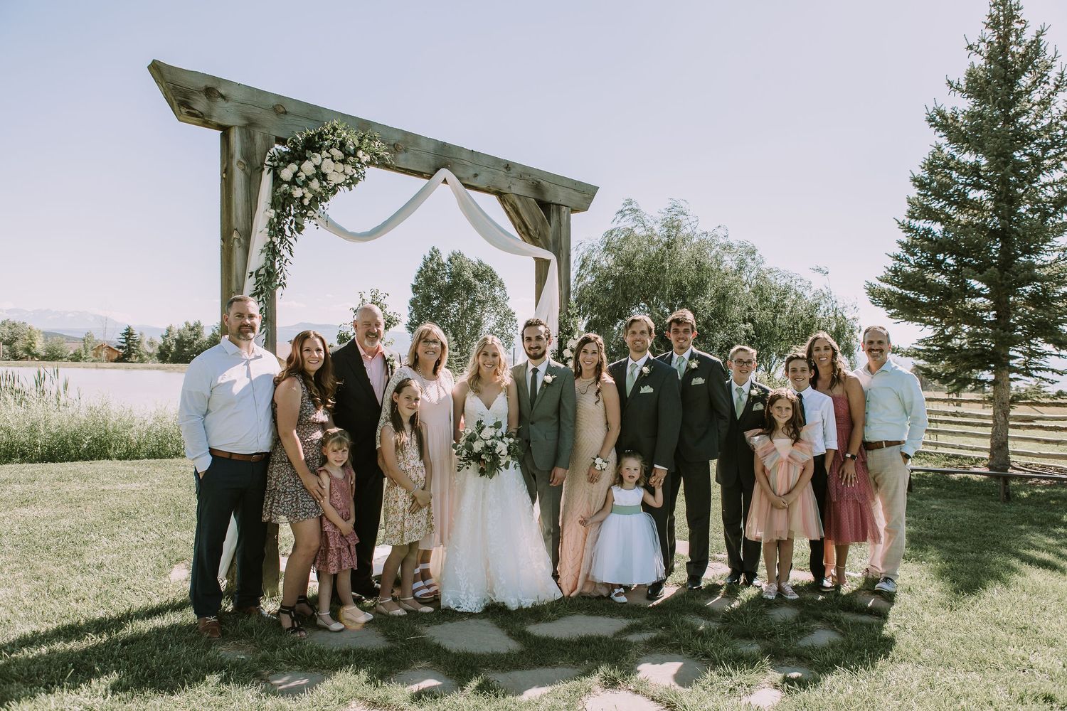 Large wedding party group photo under wooden arch in sunny outdoor garden setting.