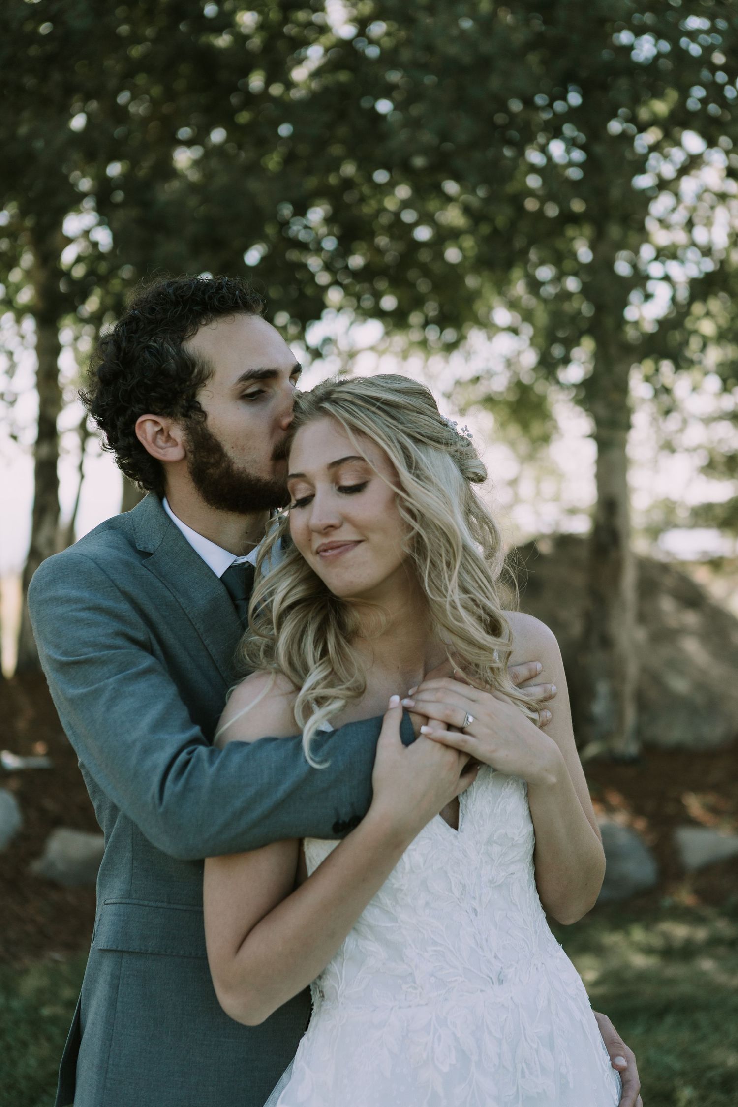 Tender moment between newlyweds as groom kisses bride's temple in outdoor setting.