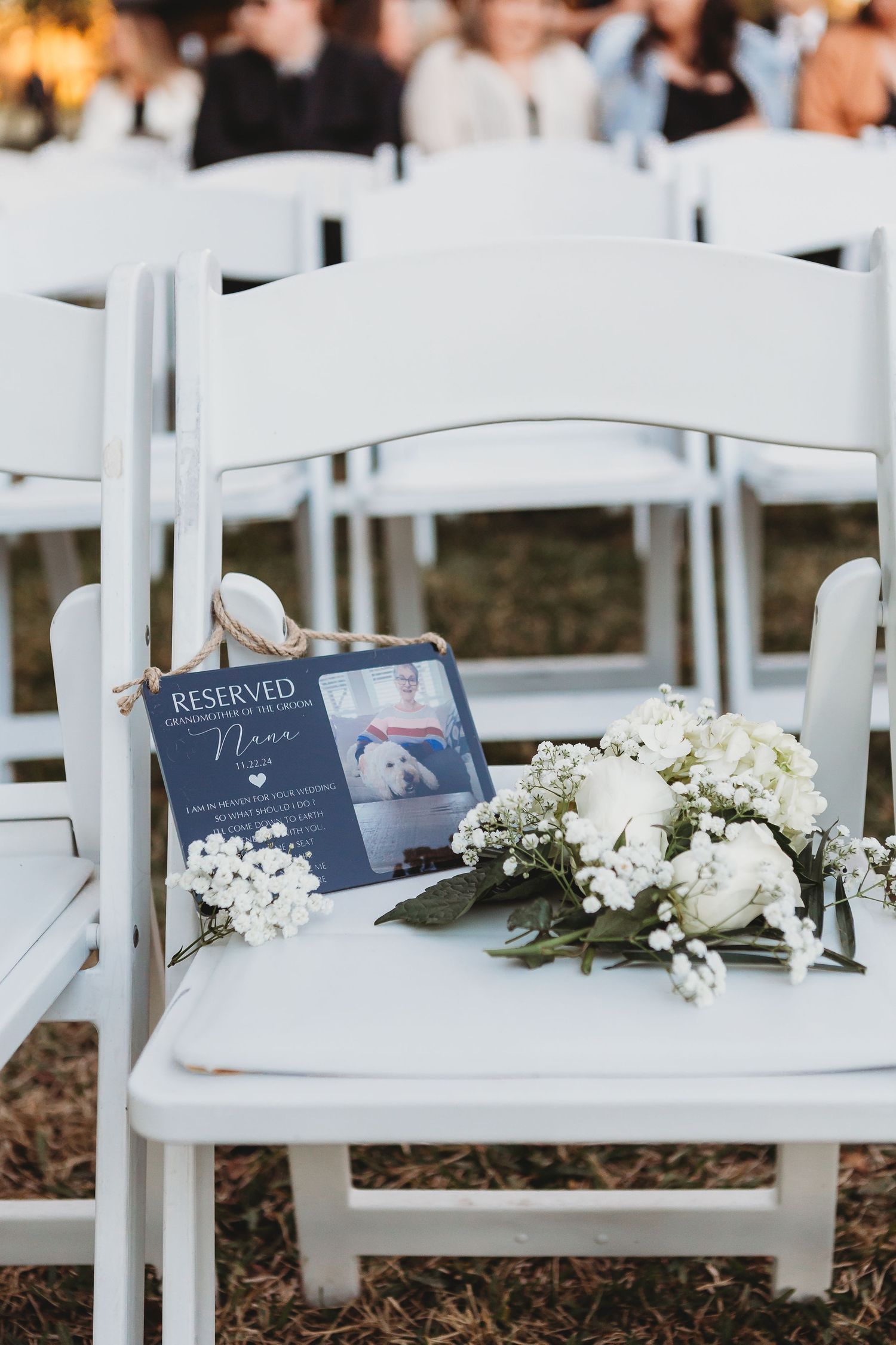 Reserved seating sign and white floral arrangement on white folding chair at outdoor ceremony.