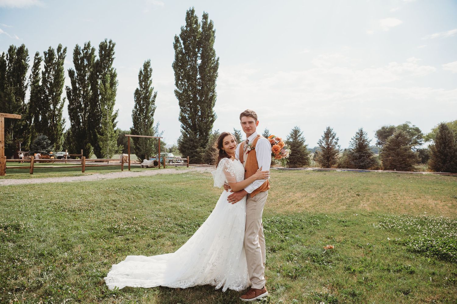 A couple in wedding attire embraces outdoors with tall poplar trees in the background.