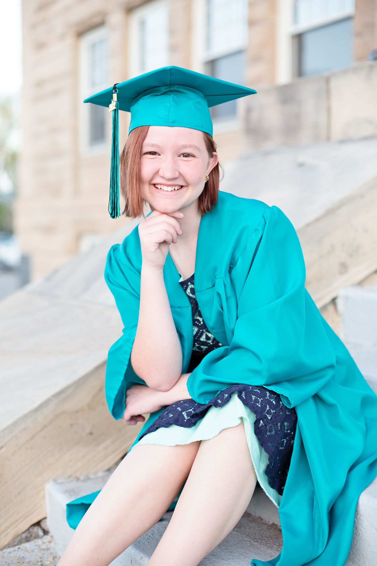 Graduate in teal cap and gown sitting on stone steps during graduation photoshoot.