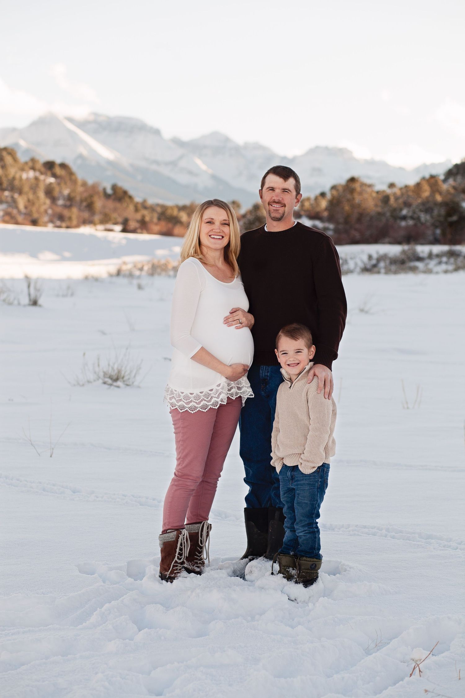Family of three poses together in snowy winter landscape with mountains in background.