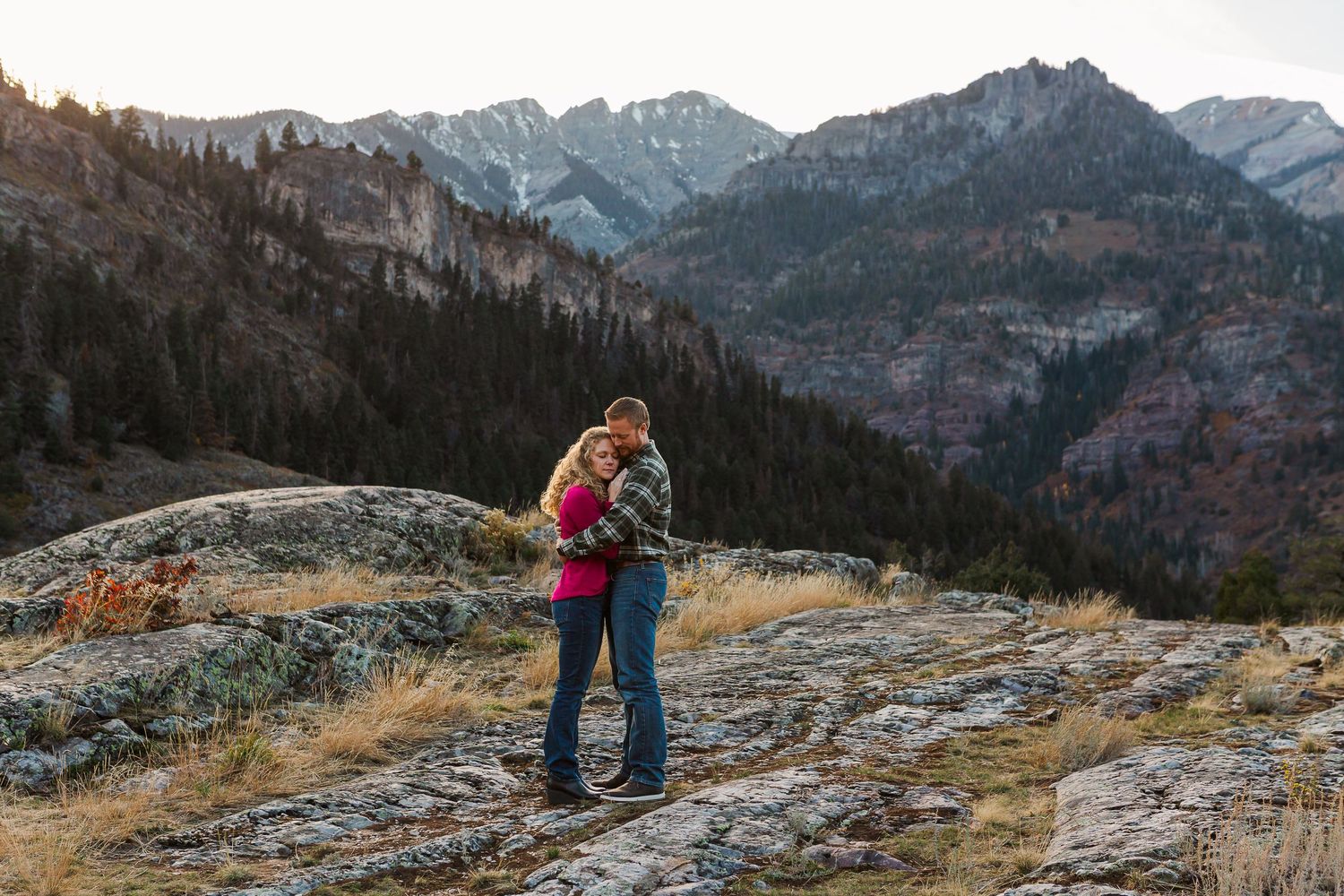 A couple shares a tender moment on a rocky mountain overlook with a panoramic valley view.