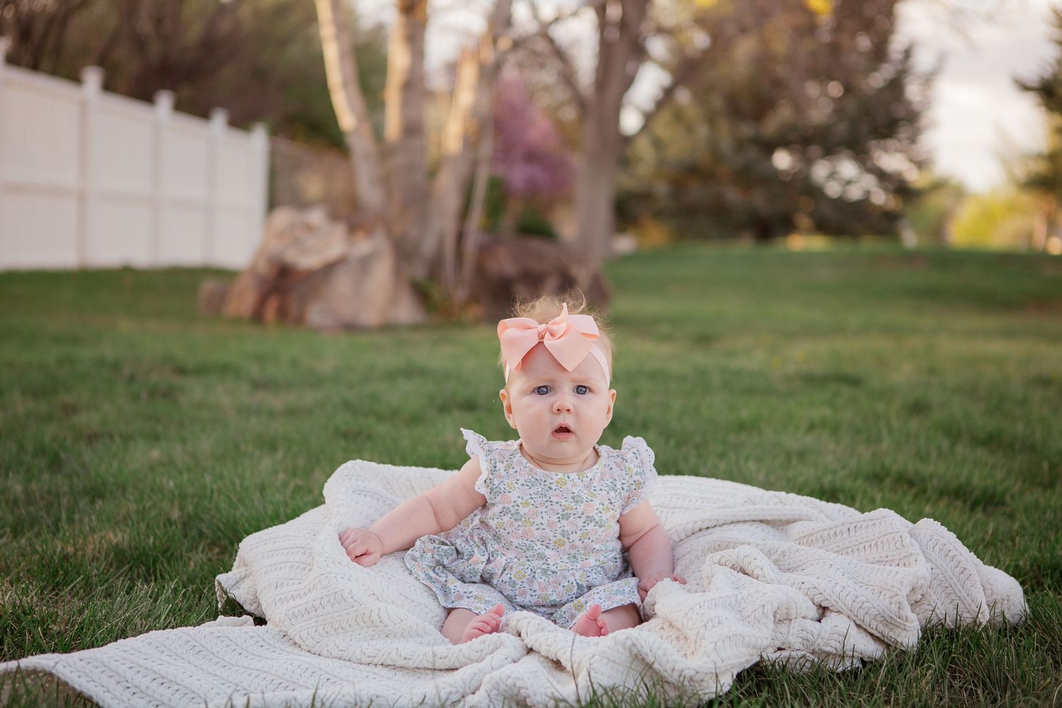 Baby in lace dress sitting on white blanket outdoors during sunset.