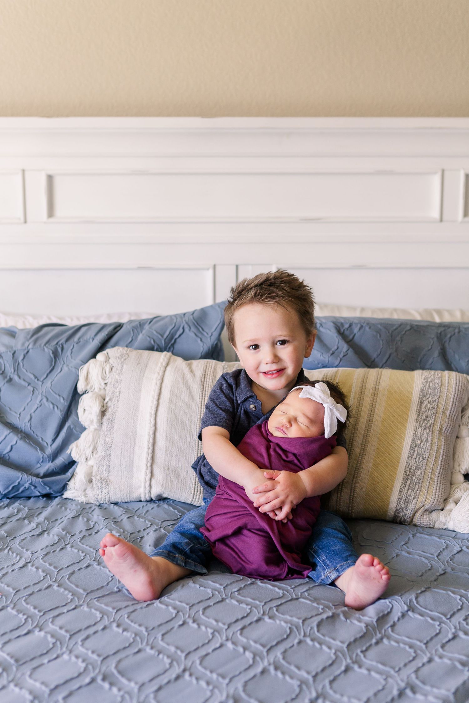 Young child holding newborn sibling while sitting on quilted gray bedding.