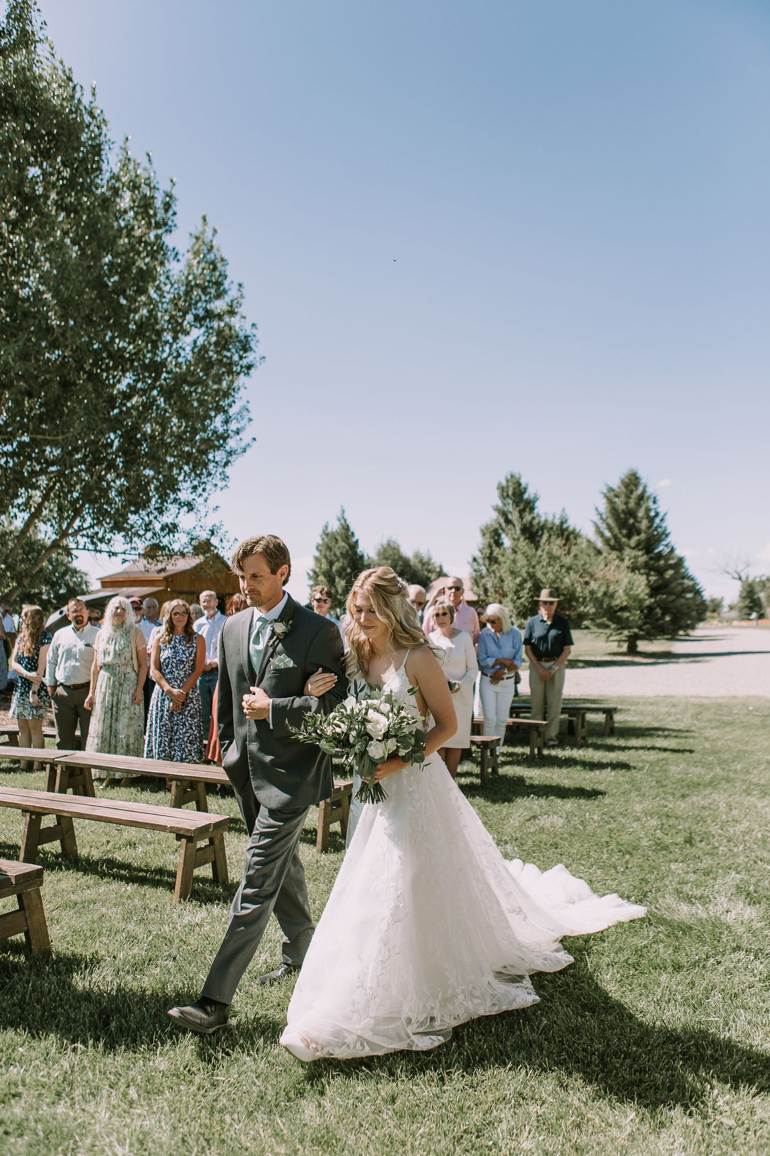 Newlyweds walking down aisle at outdoor wedding with rustic wooden benches.