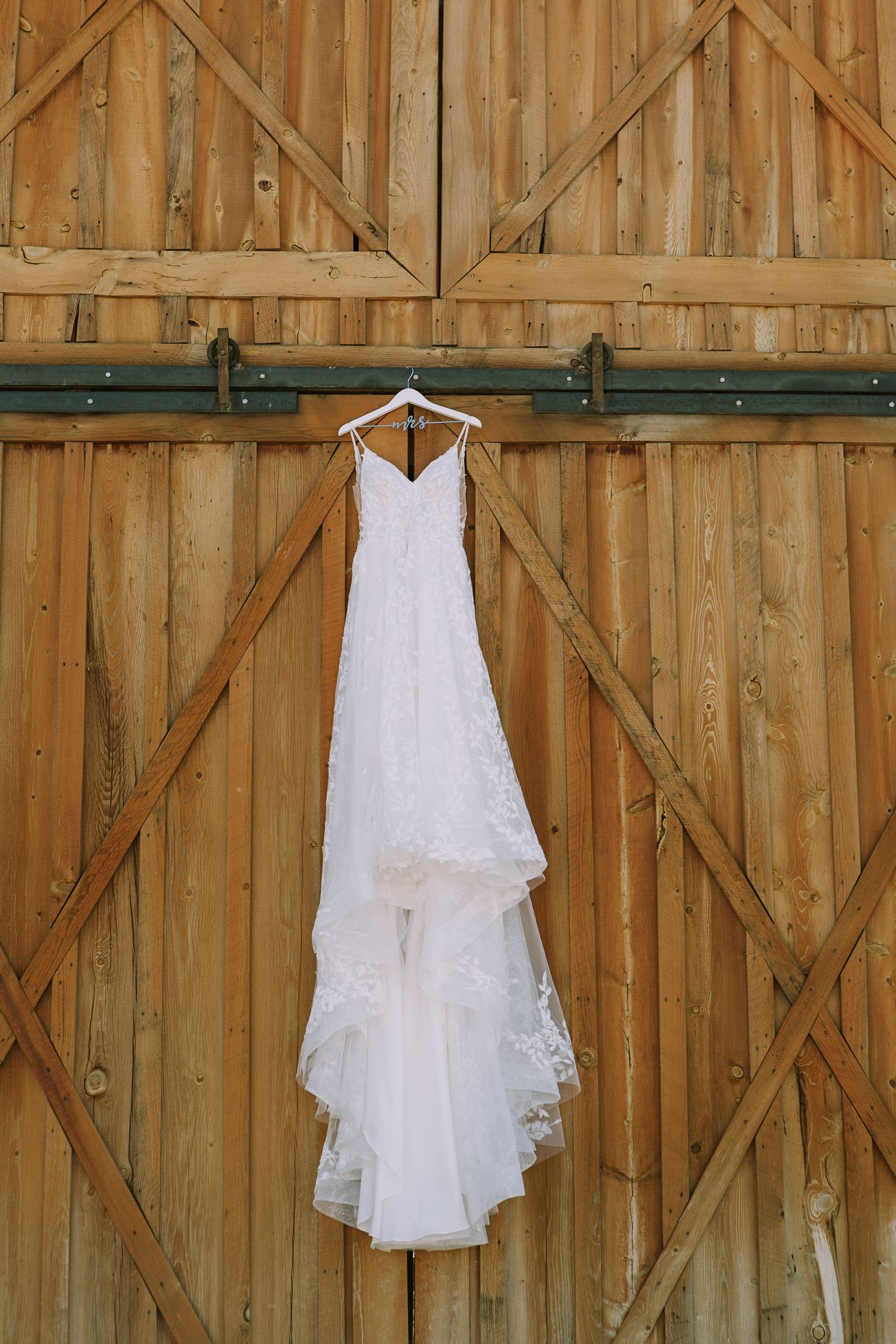 White lace wedding dress hanging against weathered wooden barn door.