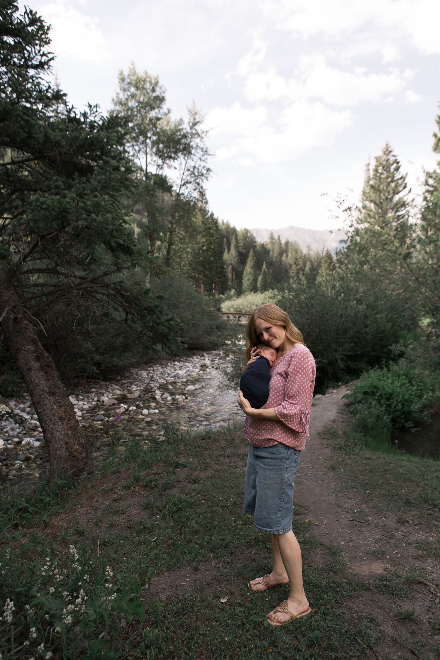 A parent holds an infant while standing on a path beside a mountain stream.