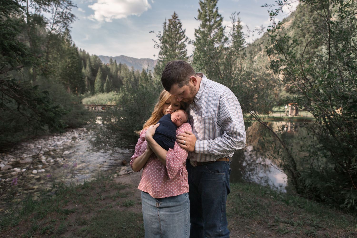 Family shares tender moment on mountain trail with stream and forest backdrop.