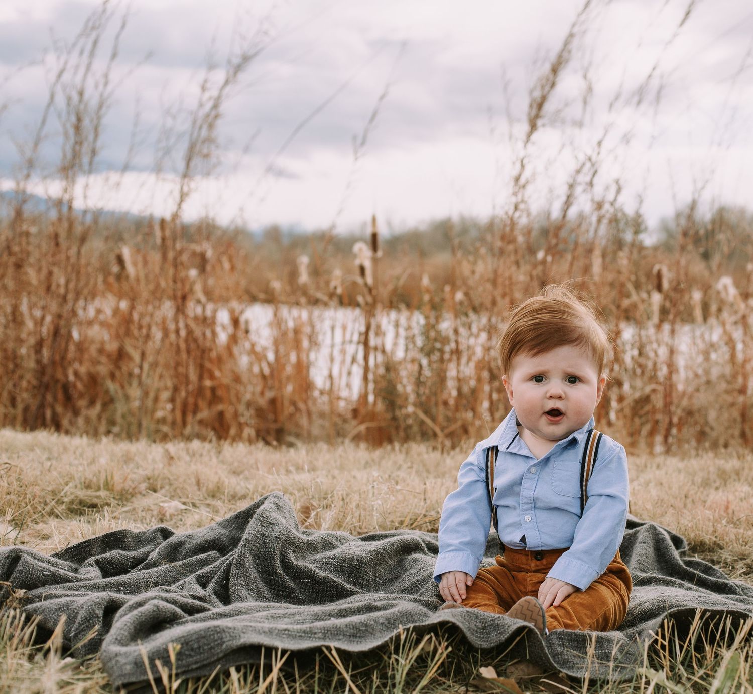 Child wearing suspenders and brown pants sits on blanket by pond with tall grasses.