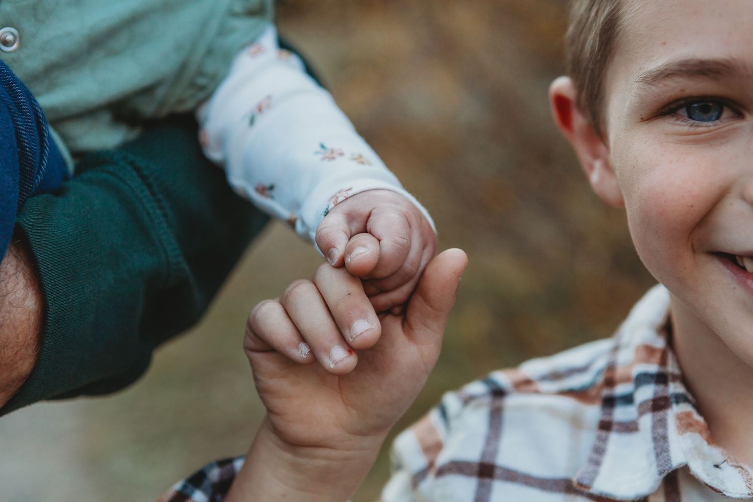 Child in plaid shirt holds hands with someone wearing light colored long sleeves in outdoor autumn setting.