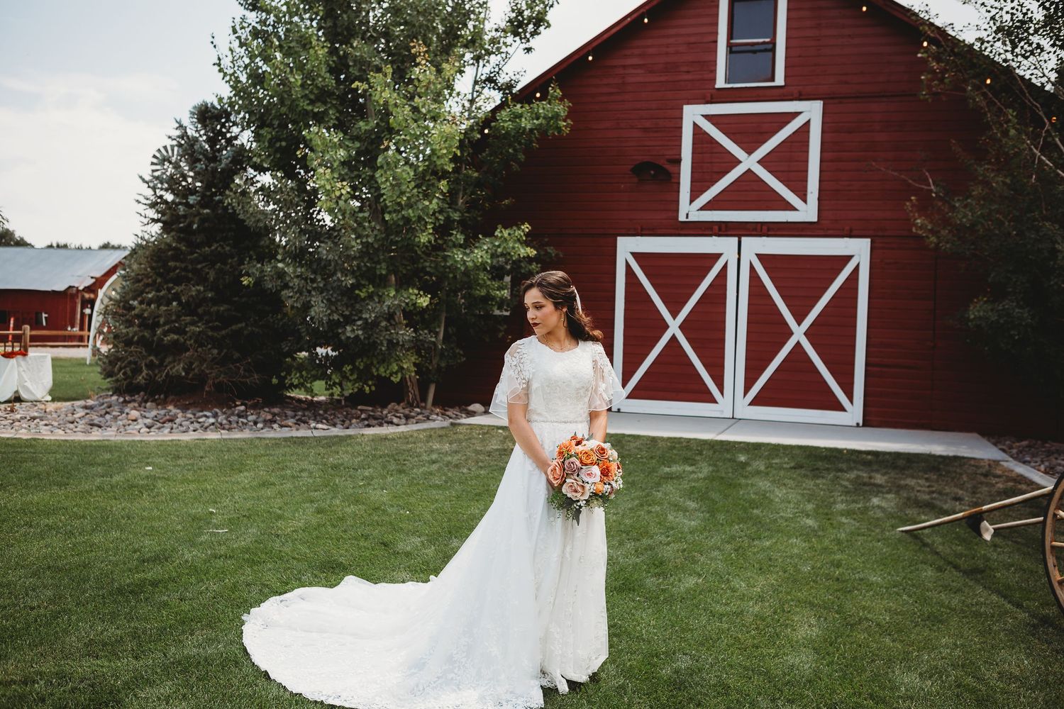 A bride in flowing white dress poses in front of red barn with white trim.