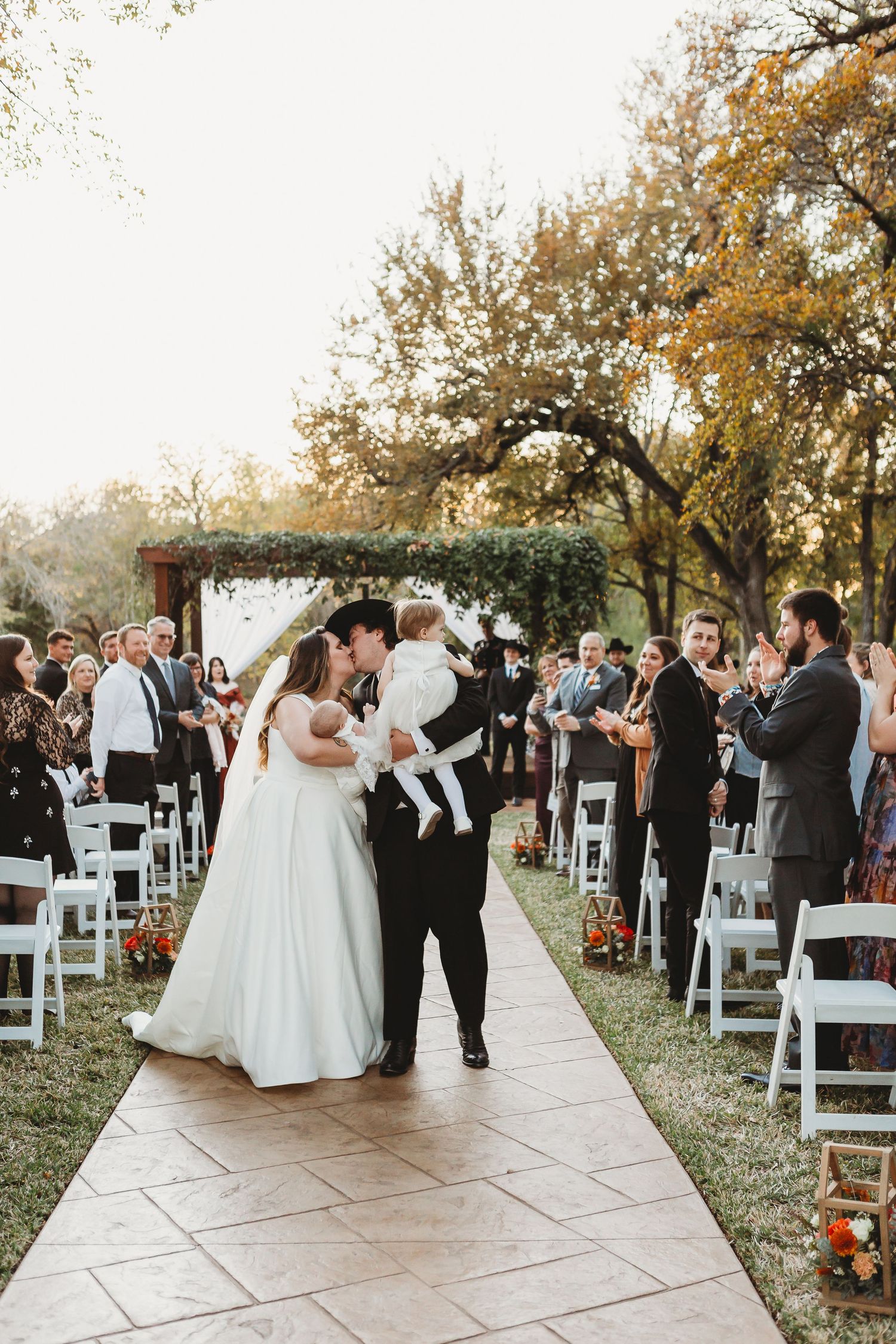 Newlyweds share first dance on stone pathway while guests watch from white chairs.