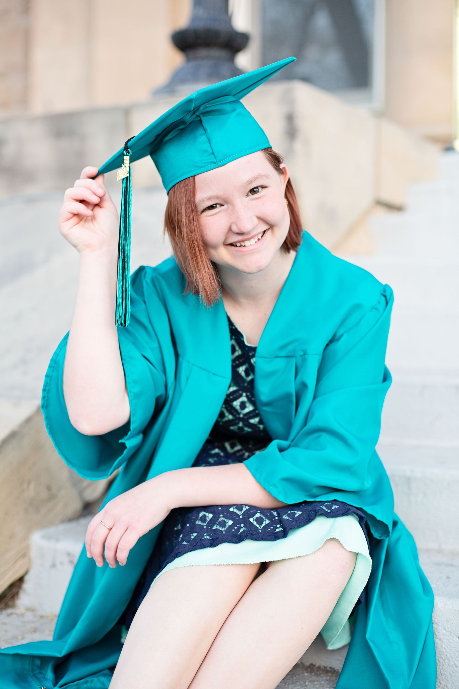 Graduate in teal graduation attire adjusts cap while sitting on stone steps.