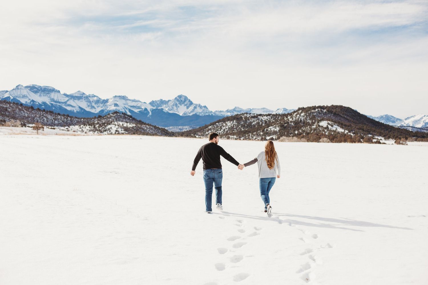 Two people run hand in hand across a snow-covered field with mountains in the distance.