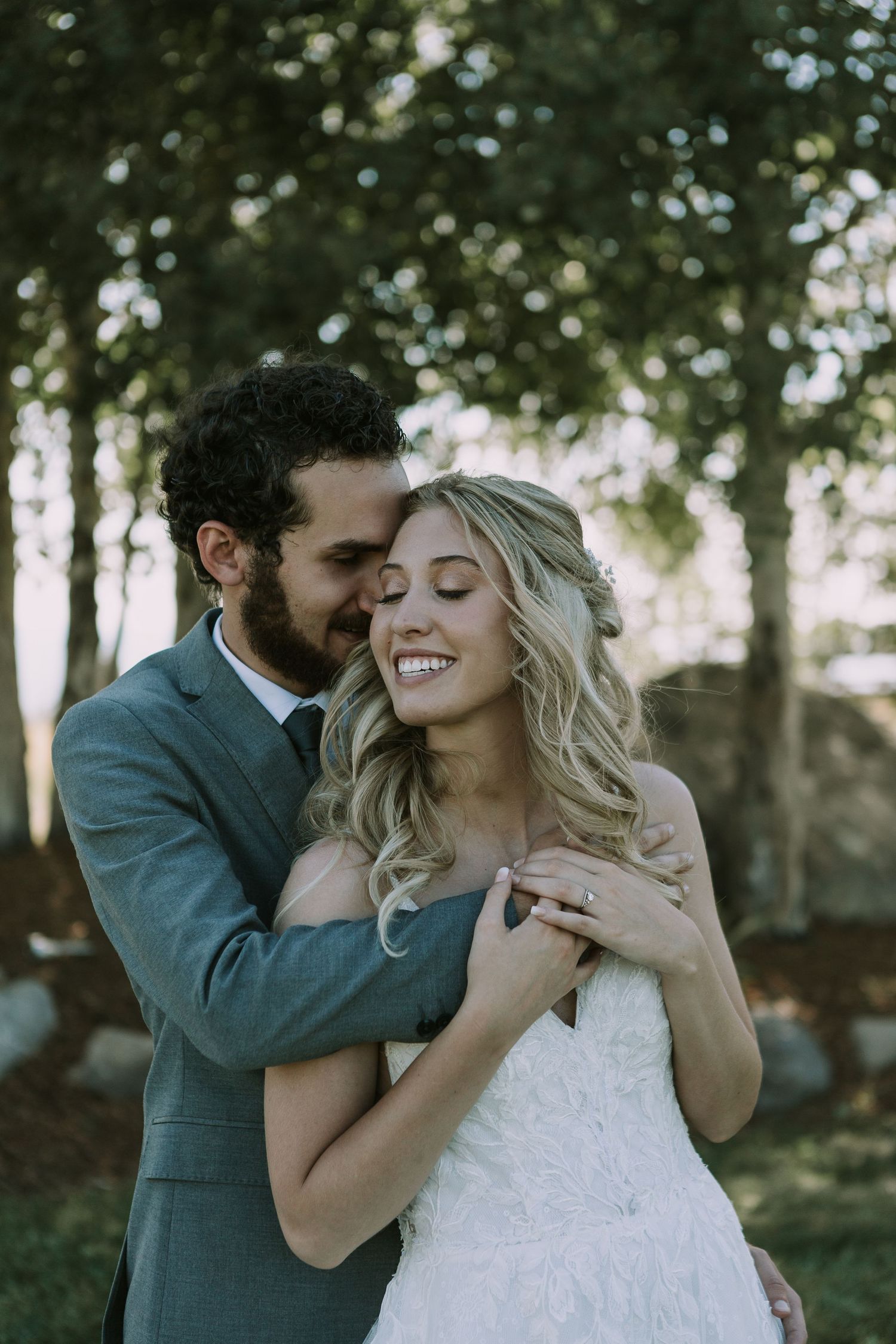 Candid wedding photo shows couple sharing intimate moment under trees with natural light.