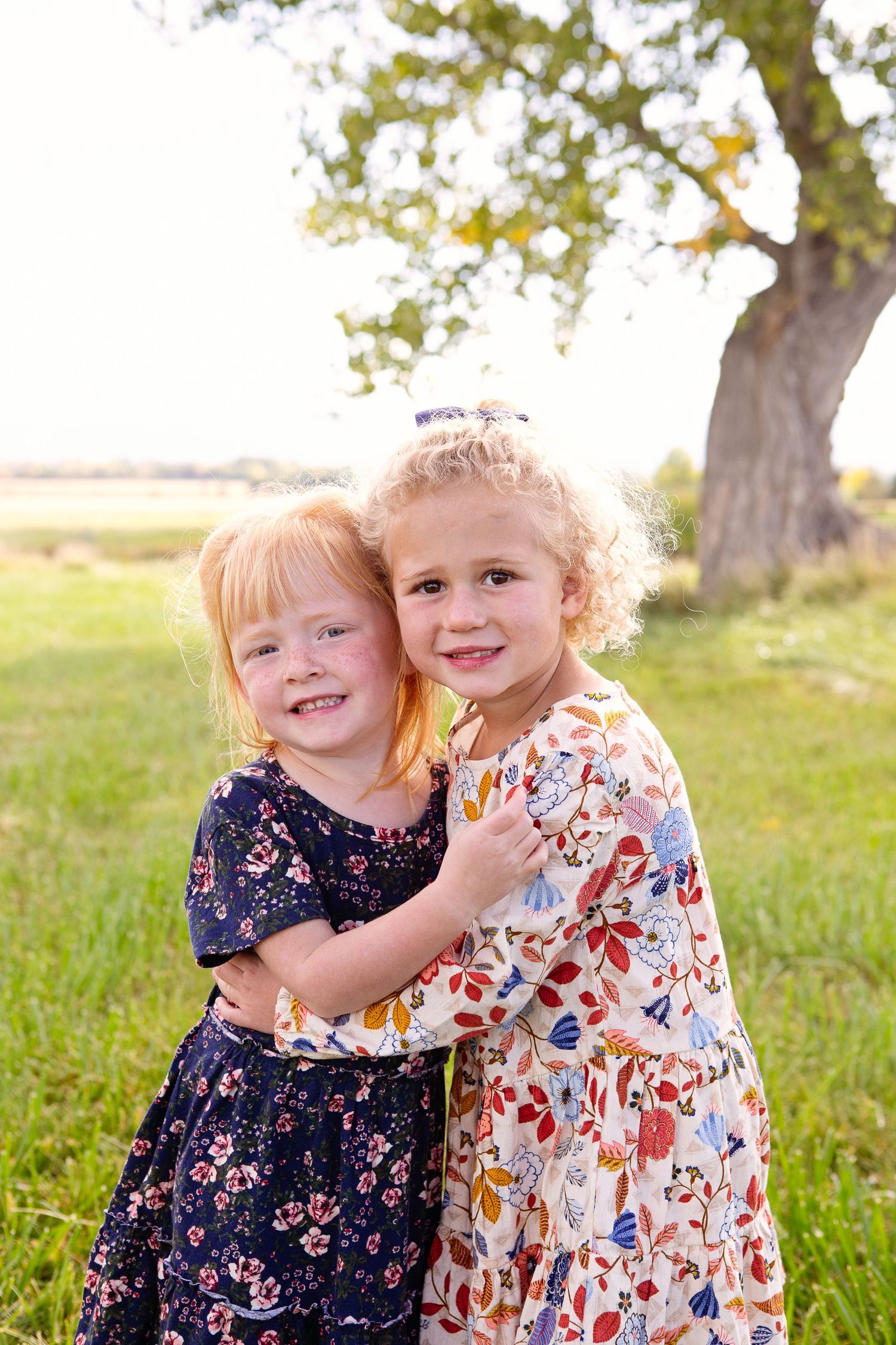 Two children wearing floral dresses embrace while standing in a sunny meadow under a tree.