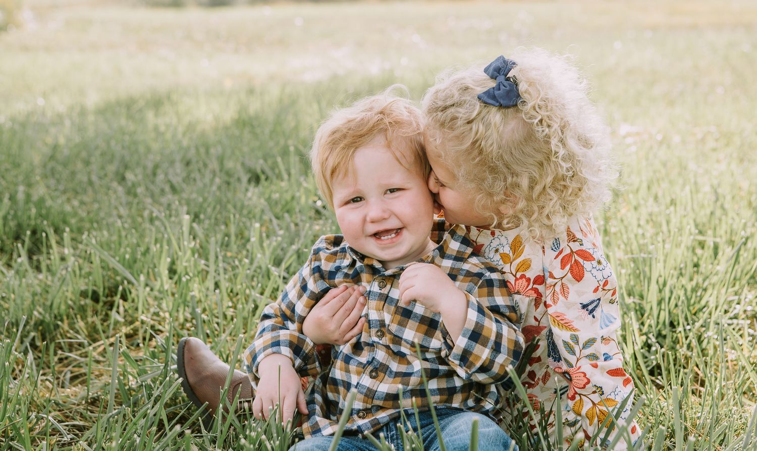 Two children share a playful moment while sitting in golden sunlit grass.