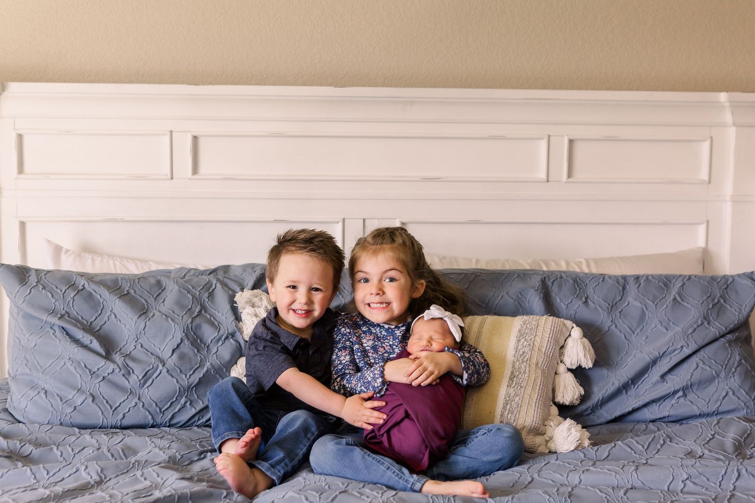 Siblings sitting on bed with gray bedding while holding newborn baby.