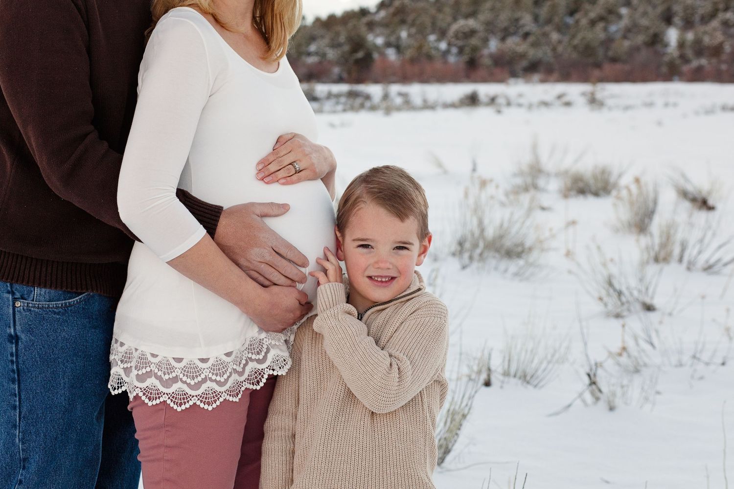 Winter maternity photo with family standing together in the snow-covered landscape.