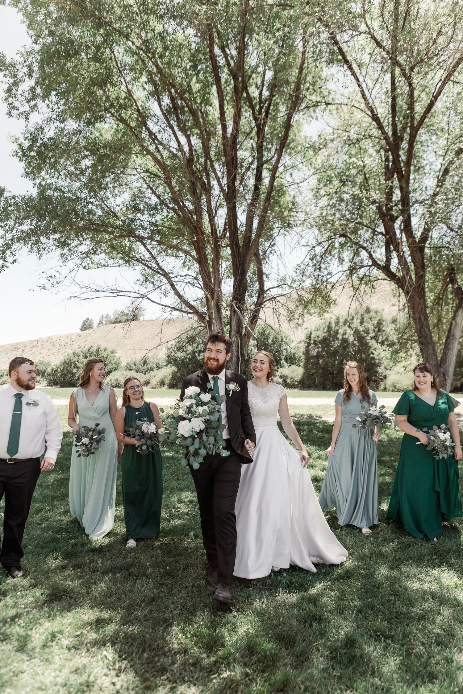 Elegant wedding party photo with mixed green bridesmaid dresses against natural backdrop.