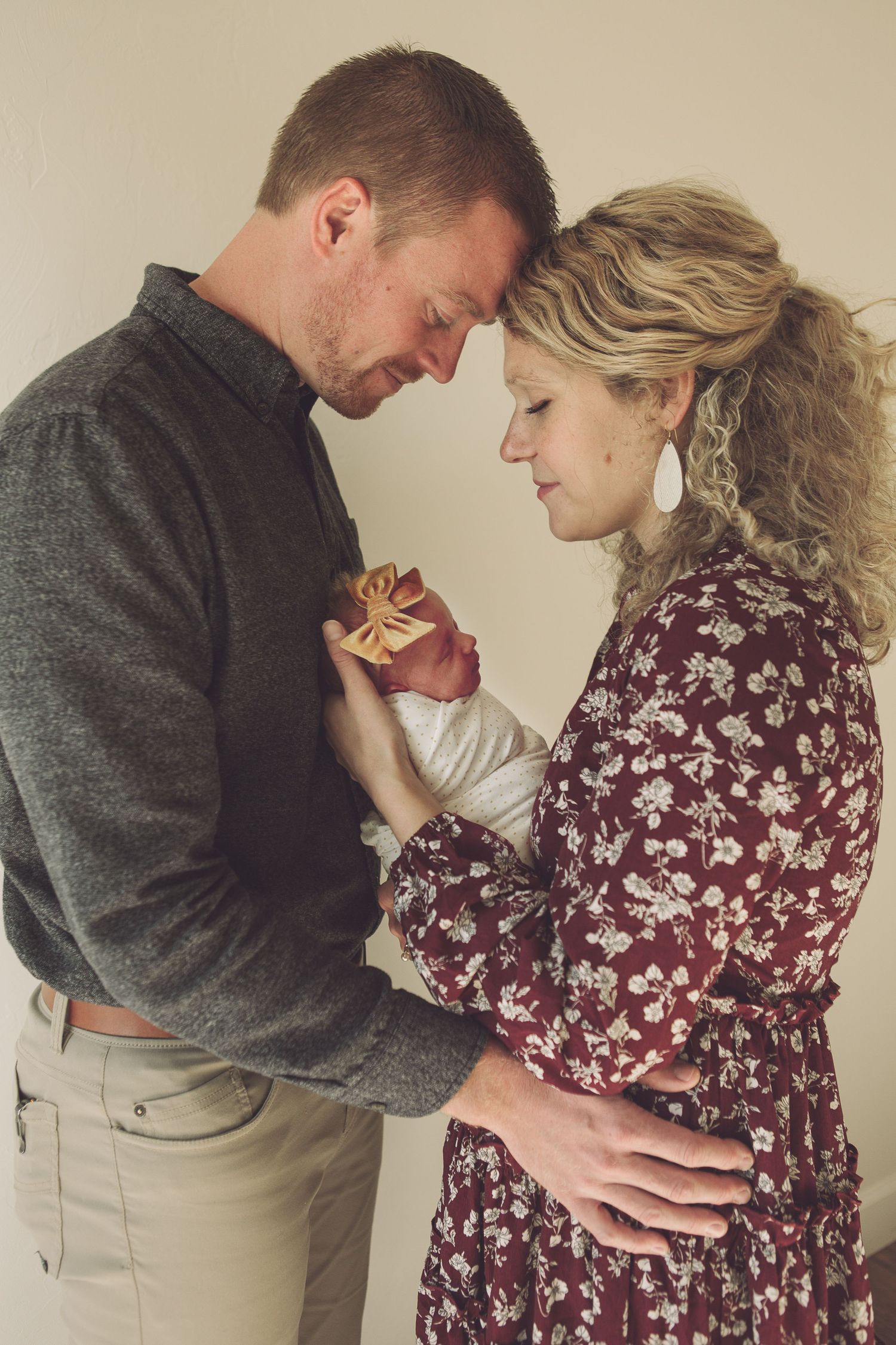 Couple in grey and floral burgundy dress share a loving moment while holding their infant.