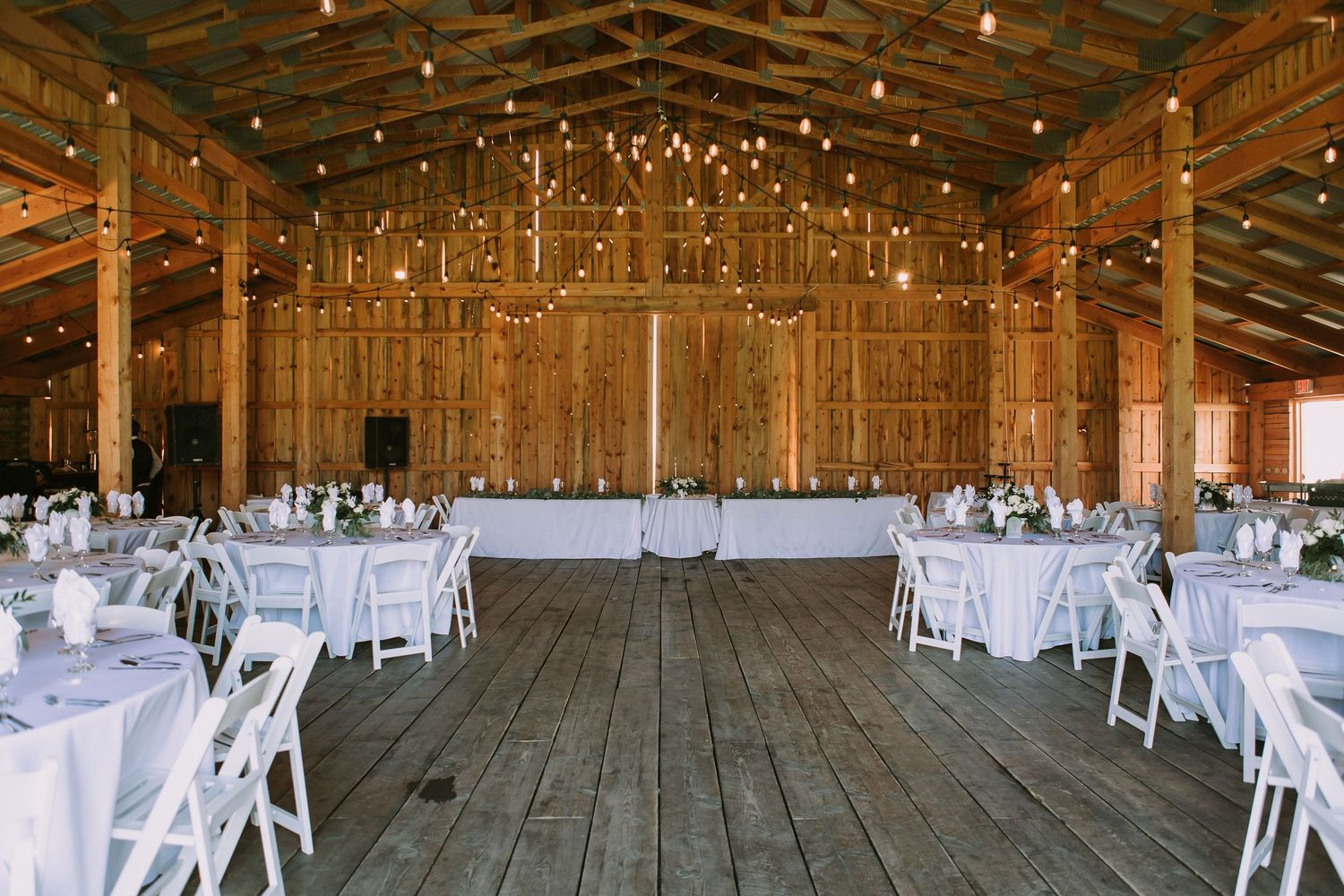 Rustic barn wedding venue with string lights, white tables, and exposed wooden beams.