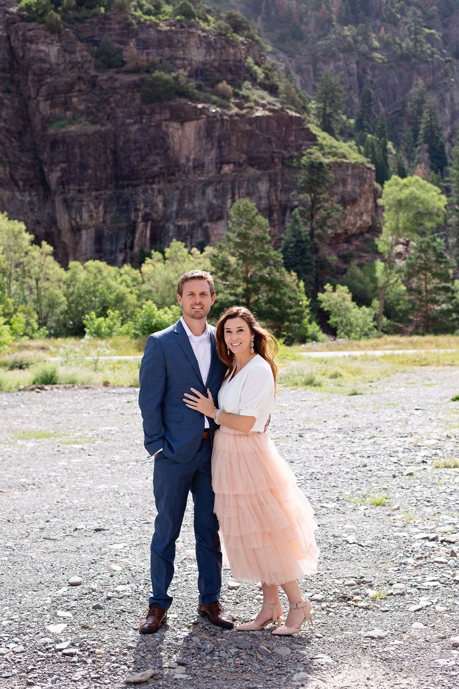 Couple embracing in formal wear against scenic mountain backdrop.