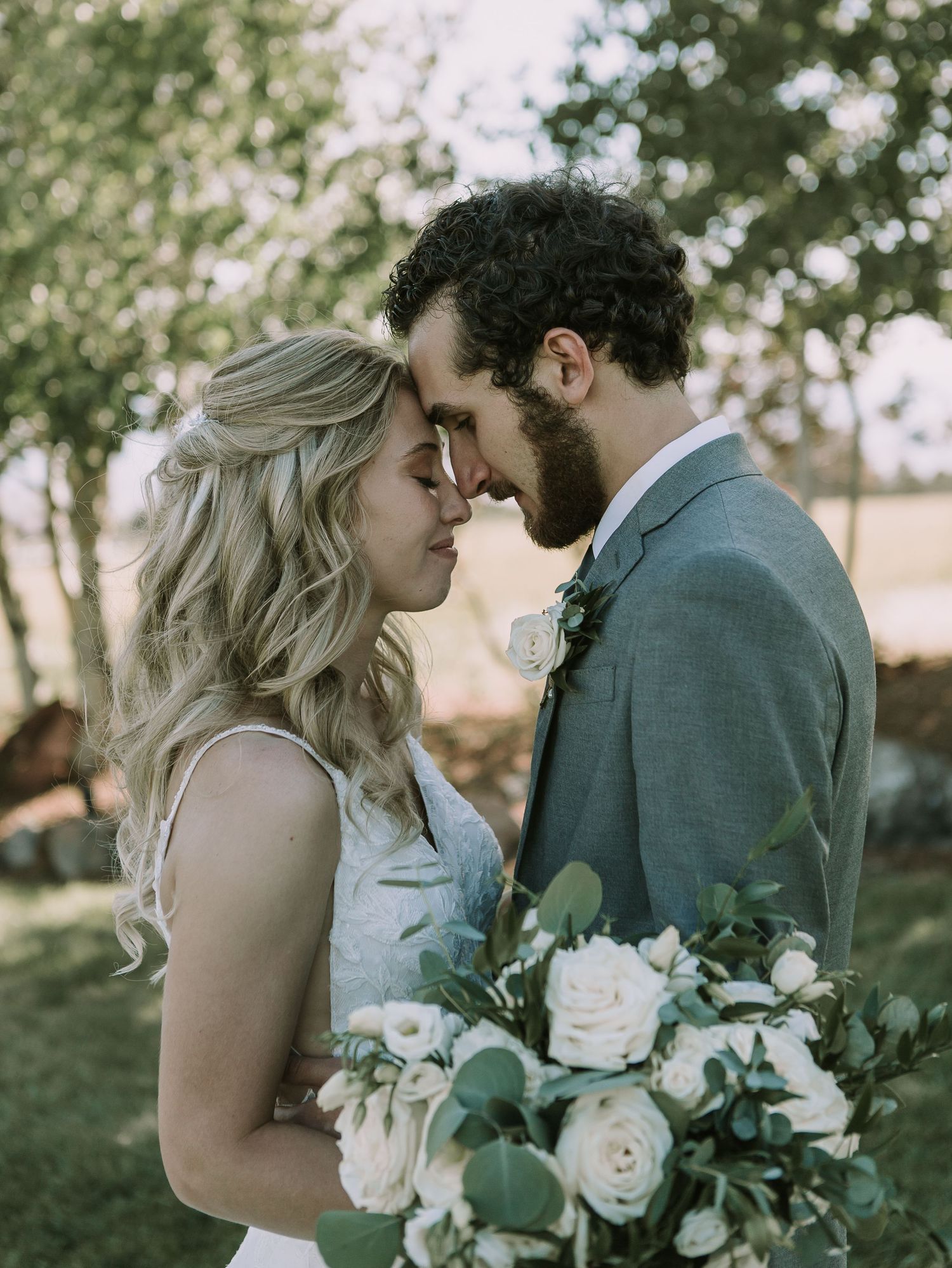 A tender forehead touch between newlyweds holding a romantic bouquet of white roses and greenery.