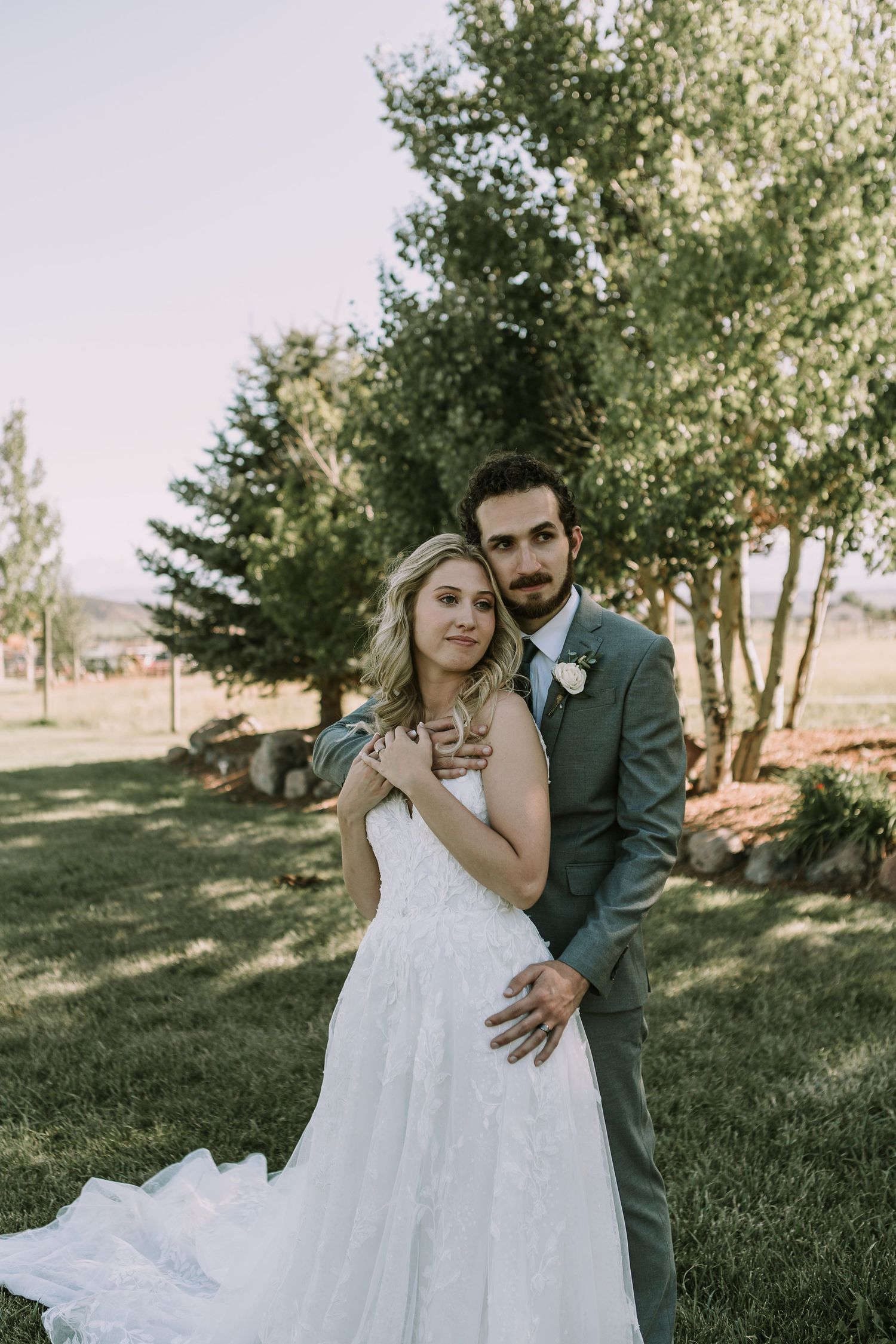 A romantic wedding portrait outdoors with a white lace gown and grey suit against a backdrop of trees.