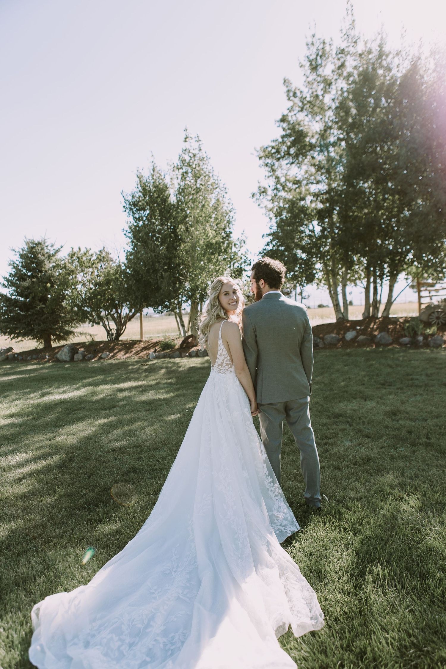 Newlyweds stand together on a grassy hill with sunlit trees, showcasing the bride's flowing lace train.