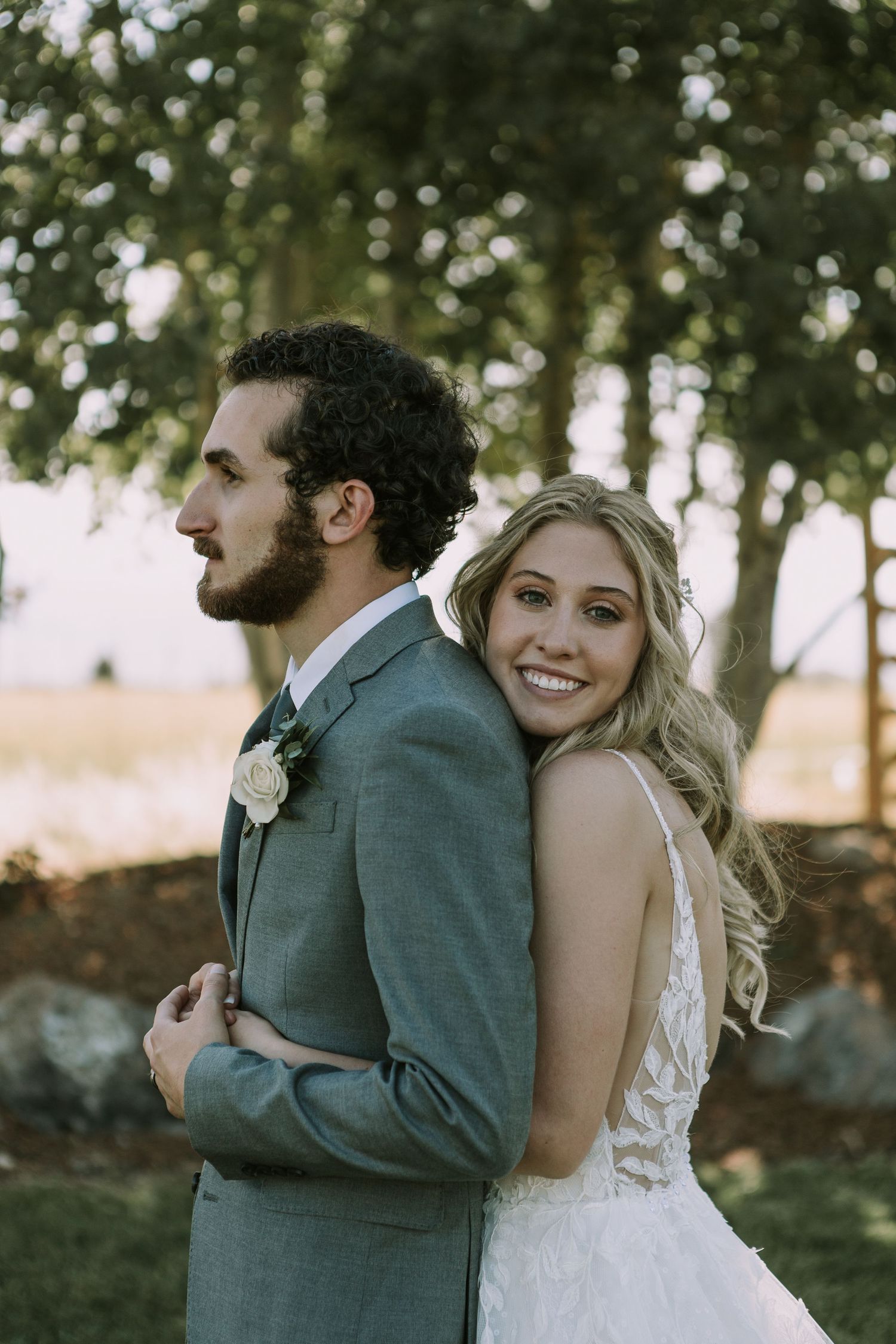 Bride in lace dress hugs groom from behind as they share a candid moment outdoors.