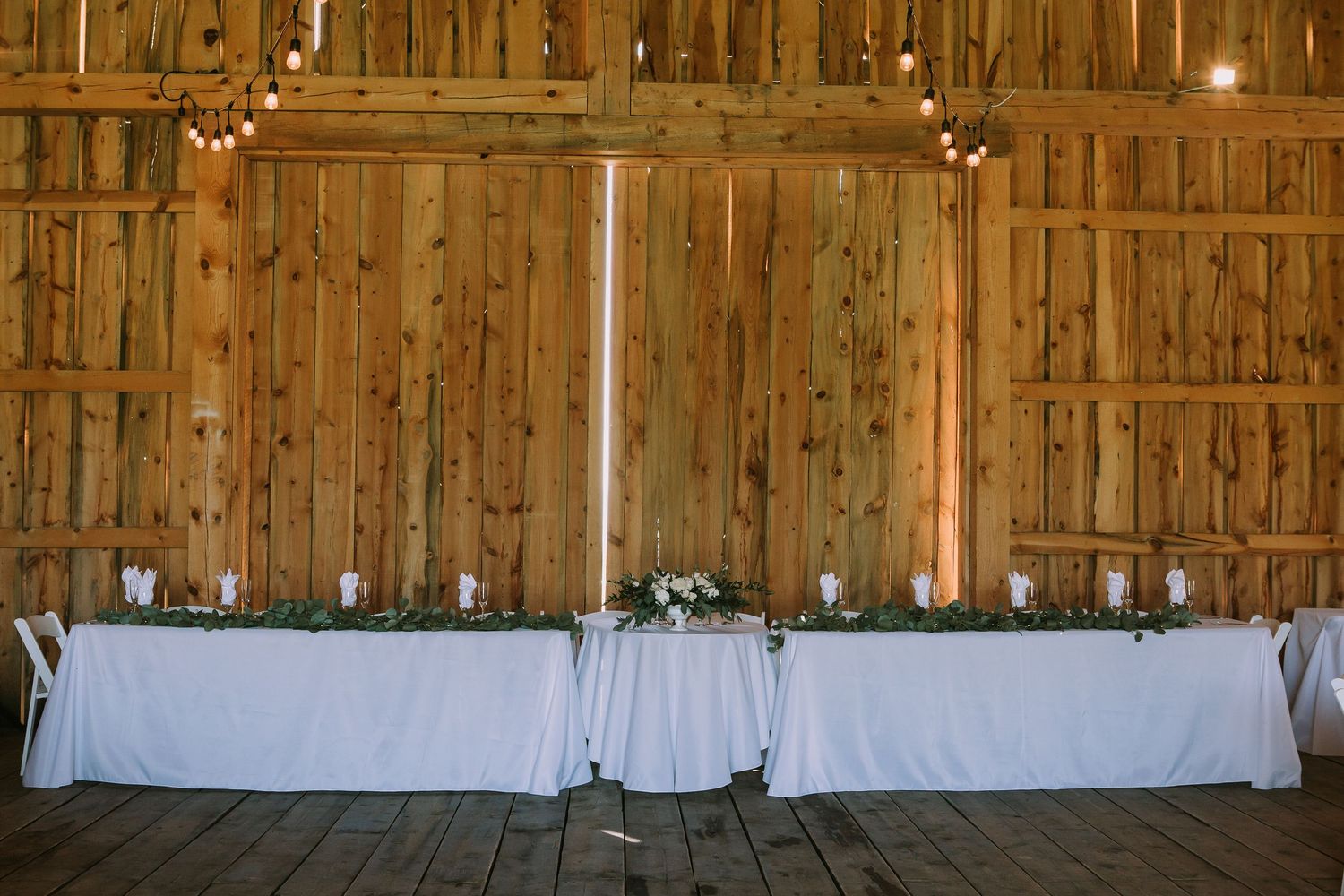 Long white-clothed tables against rustic barn wall with simple floral decor and candles.